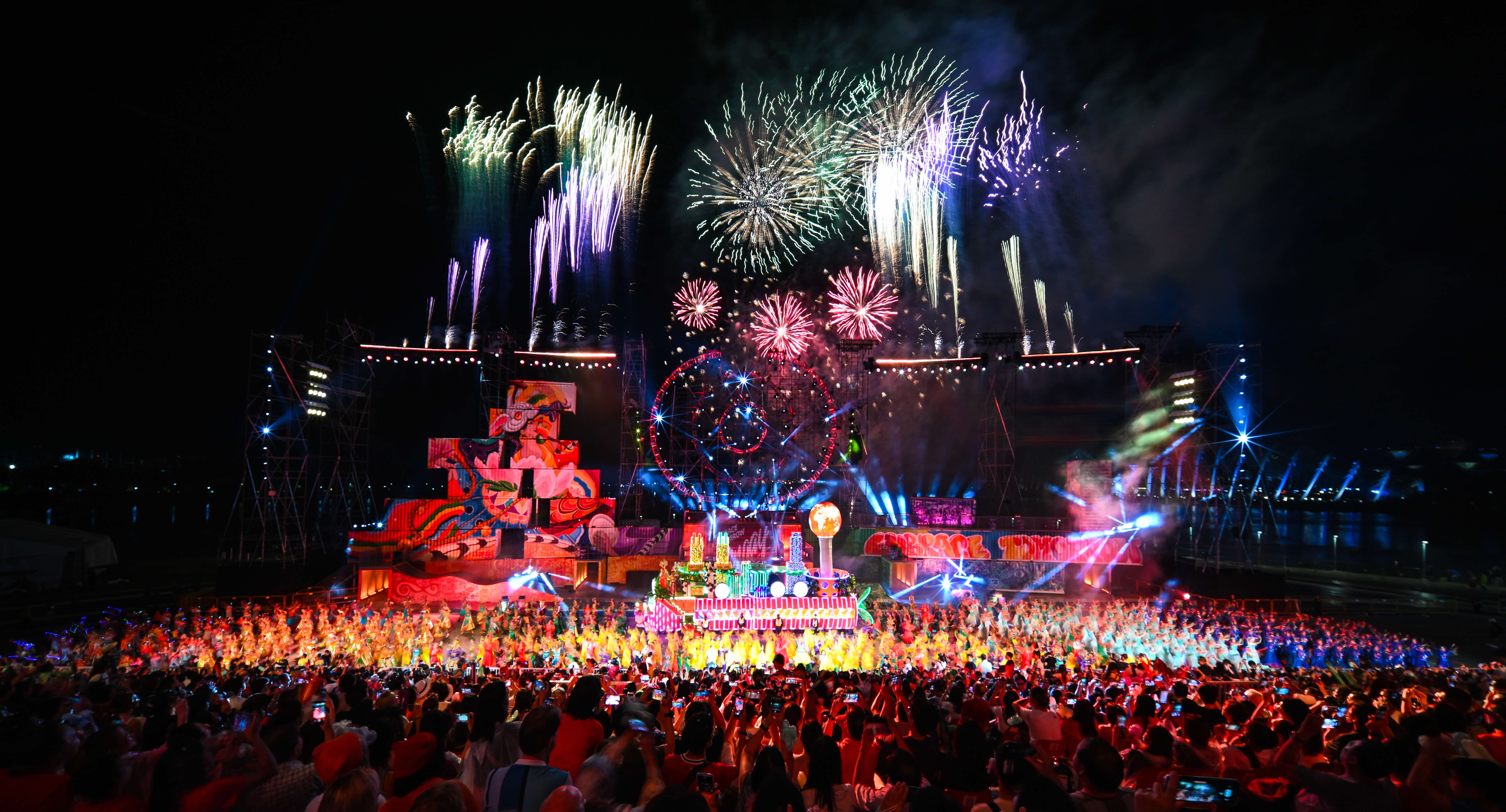 Fireworks explode over a colorful, crowded stage at night with vibrant lights and numerous performers entertaining the audience.